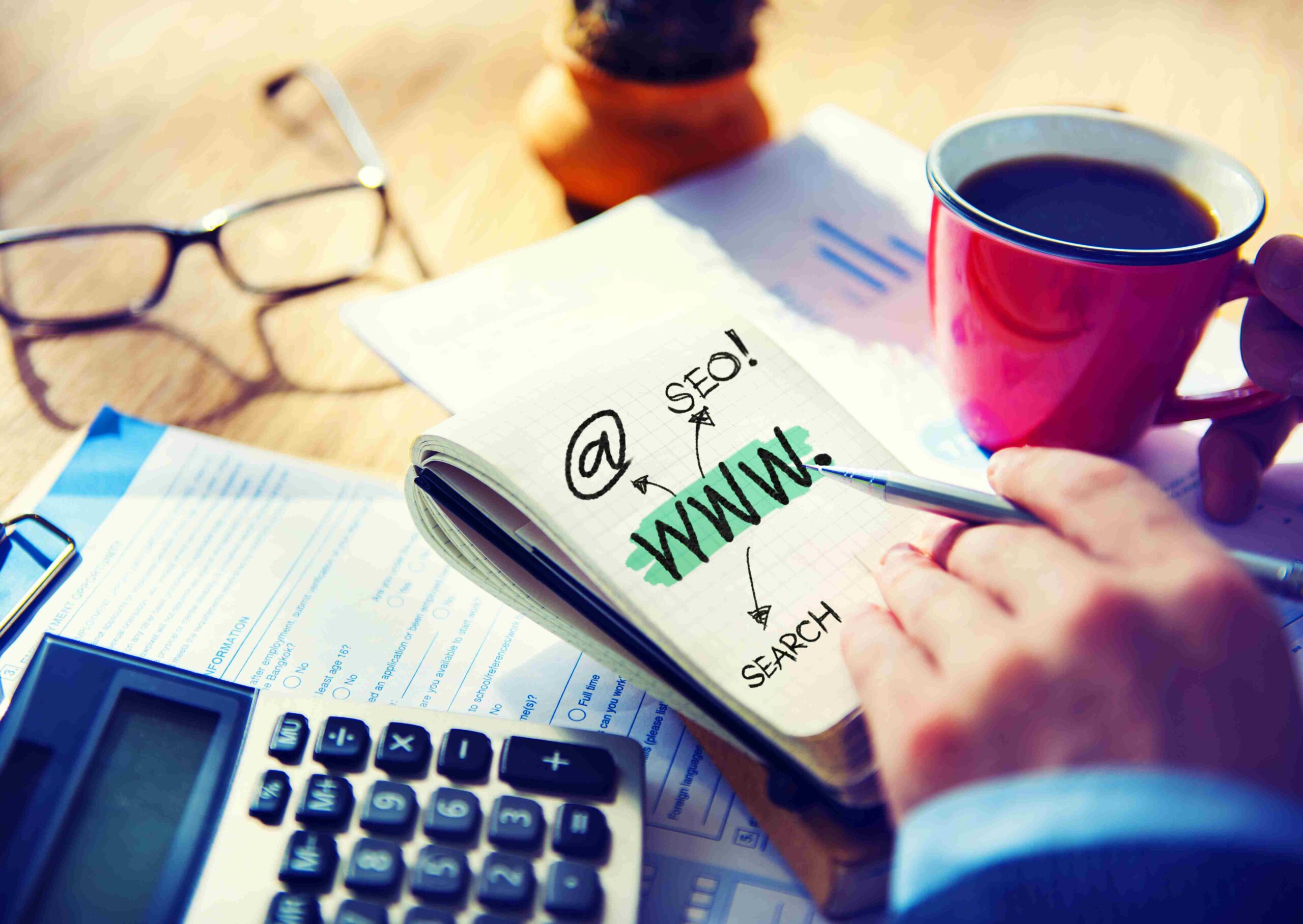 Beležnica sa napisanim SEO i WWW pored kalkulatora i šolje kafe što simbolizuje planiranje online strategije